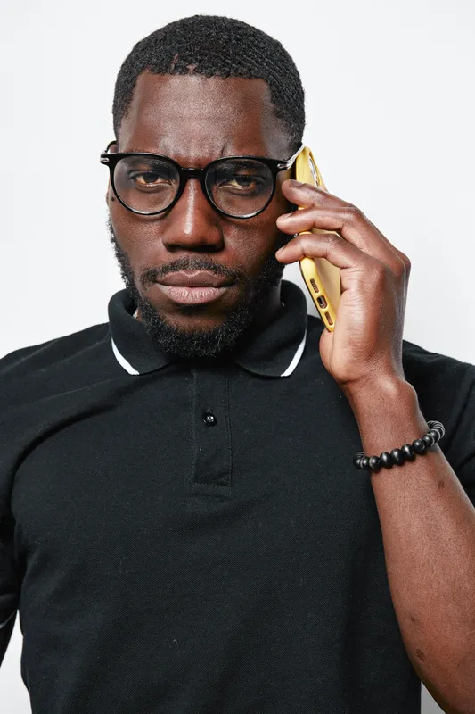 African man with dark skin wearing glasses and black polo shirt talking on smartphone with serious expression. Closeup portrait of confident black male using mobile phone indoors.