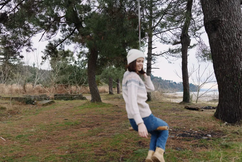Woman in a cozy winter sweater and beanie enjoys a playful moment outdoors, standing near tall trees by a calm lake with grassy patches and a light breeze, showcasing casual fashion and natural