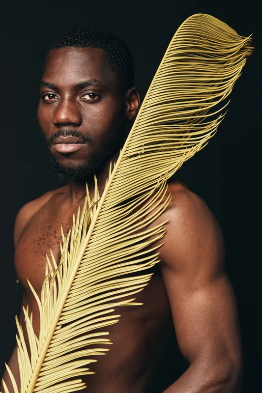 African man with dark skin and beard holding large dried palm leaf in creative conceptual visual style on black background. Artistic portrait with natural texture and expression.