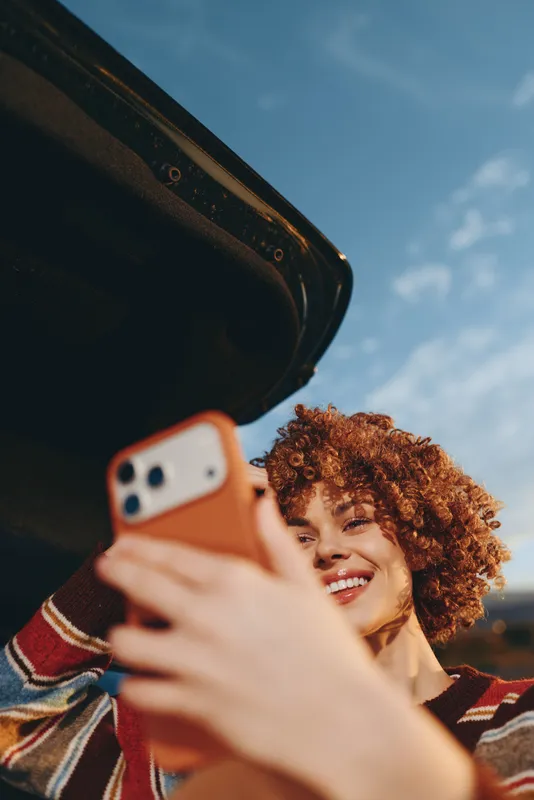 Woman takes selfie with phone outdoors, smiling with curly hair, lifestyle portrait against blue sky, close view of face and held device, bright cheerful mood in natural light.