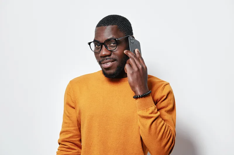 African American man with dark skin wearing glasses and orange sweater talking on smartphone with smile isolated on white background. Casual communication and technology concept.
