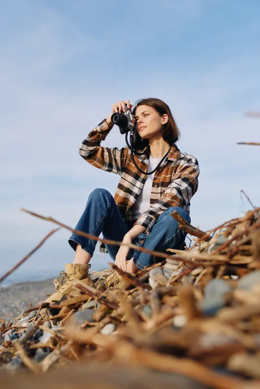 Woman photographer crouches on a rocky shore with a camera, wearing a plaid shirt and jeans, capturing coastal scenery and natural light in an outdoor moment in the open air.