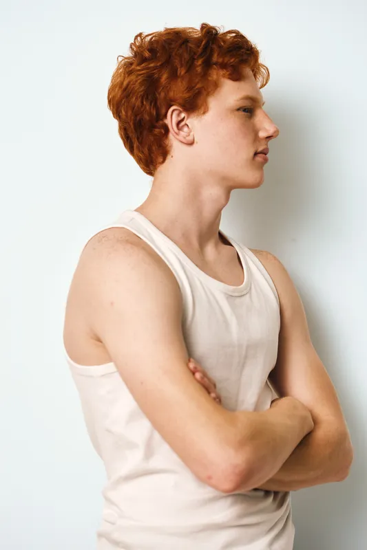 Young man with red hair and fair skin standing with arms crossed in white tank top against plain background. Profile portrait showing serious expression, casual style and youth.