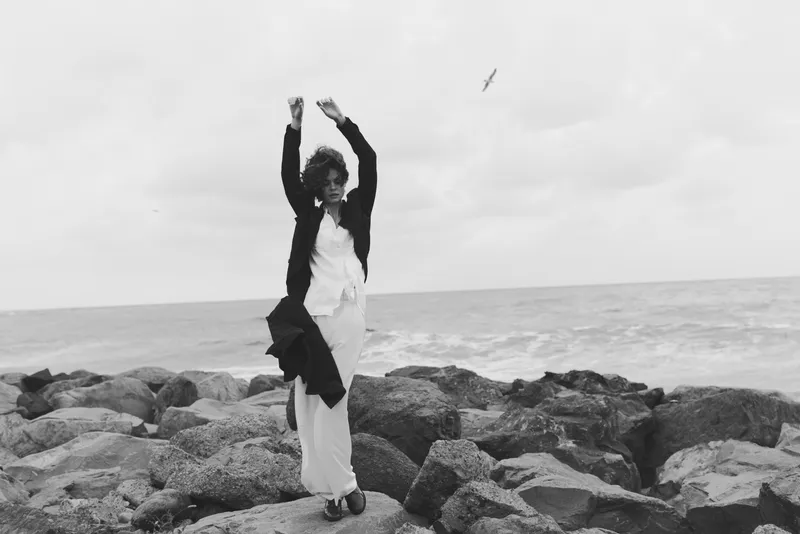 Woman dancing on rocks by the sea, jumping with arms raised, wind sweeping the scene, black and white fashion mood, dynamic coastal motion