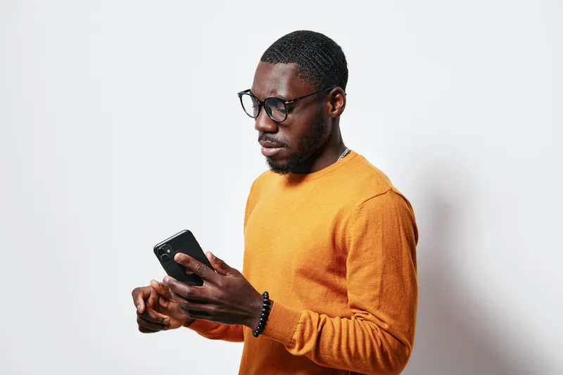 Black man with dark skin wearing glasses and orange sweater using smartphone for communication and technology on white background in studio portrait.