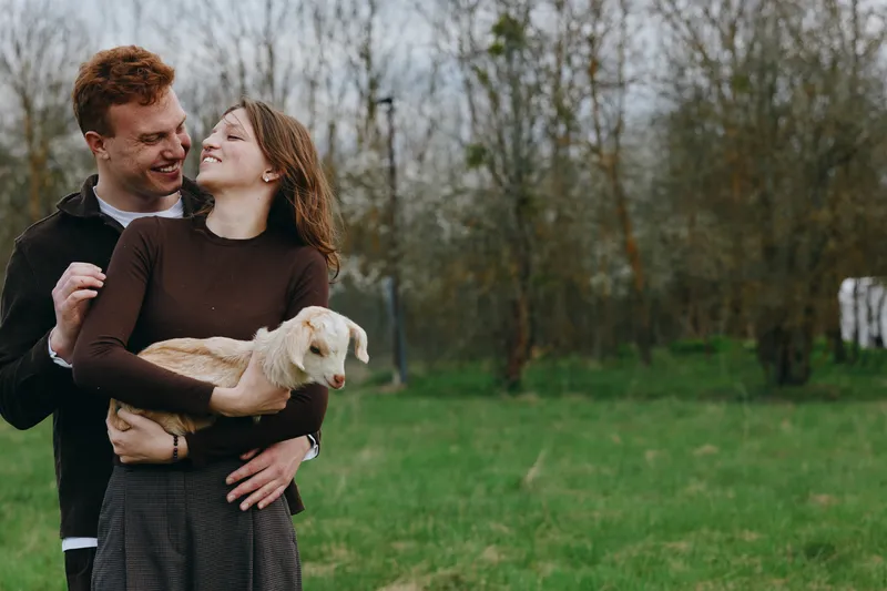 Goat kid, couple, outdoors, nature, meadow, field, grass, cheerful couple outdoors embracing a goat kid in a sunlit meadow with natural colors, candid mood, warm connection, lifestyle moment