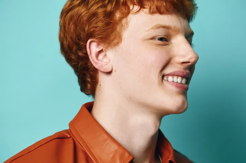 Young man with red hair smiling in profile wearing orange leather jacket with blue background for portrait fashion and happy lifestyle concept.