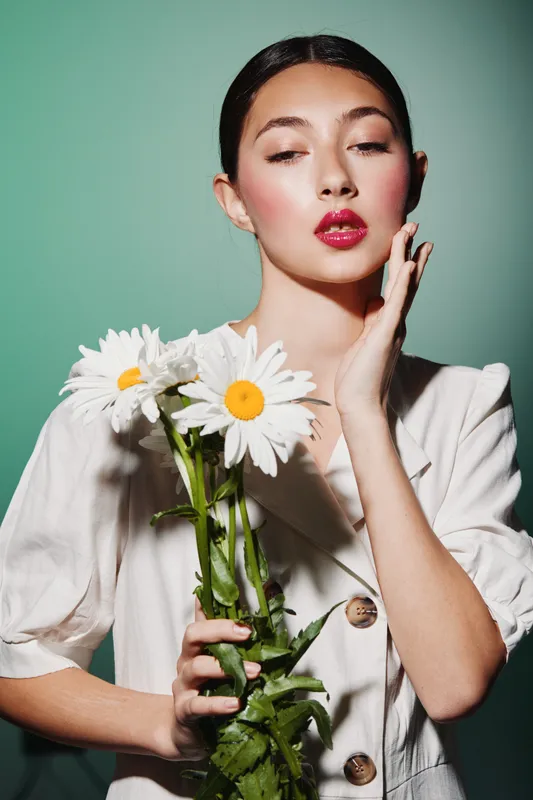 Young stylish woman in white holding white daisies looks thoughtfully into the camera. Soft makeup and glossy red lips highlight her beauty on pale teal background. Creative fashion thoughts concept.