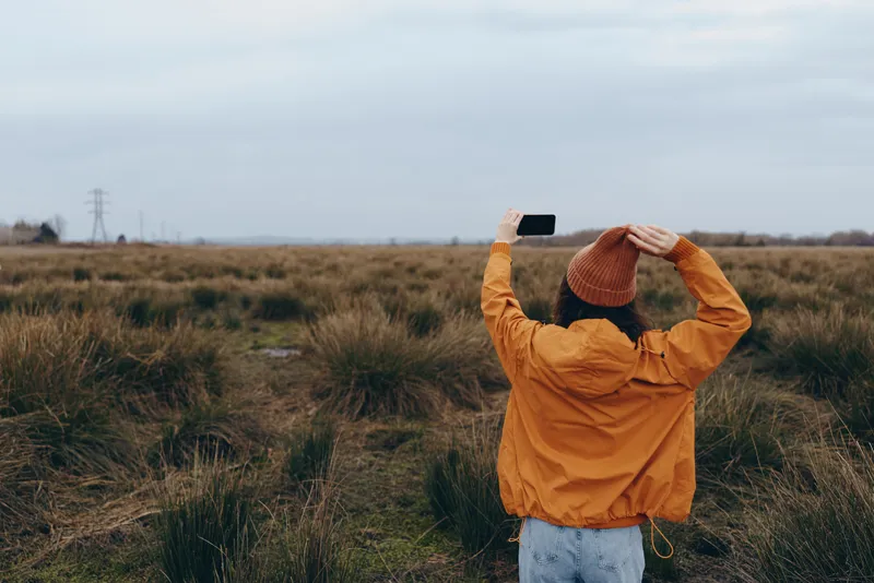 Woman lifestyle in autumn field outdoors with orange jacket and hat arms raised celebrating freedom and fresh air standing on rural path under cloudy sky