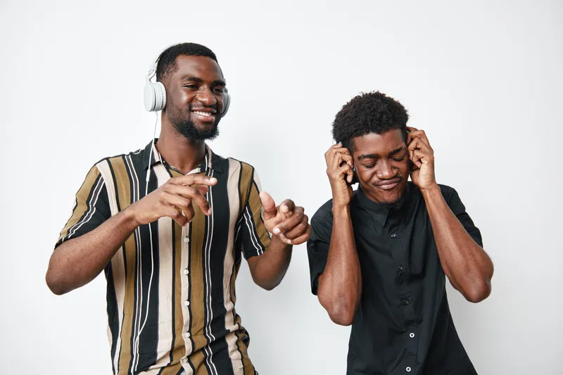 Two African American men with dark skin wearing headphones enjoying music together, smiling and dancing in casual clothes on white background in studio setting.