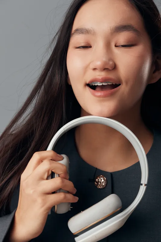 Young woman with braces holding white headphones smiling with closed eyes indoors. Female, music, technology and lifestyle concept with long hair and casual dark top on neutral background.