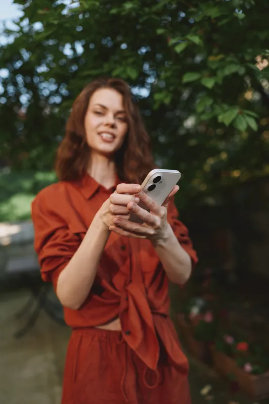 Smiling woman with wavy hair holding a smartphone for a selfie in a leafy outdoor setting. Casual fashion and carefree lifestyle conveyed through warm natural light
