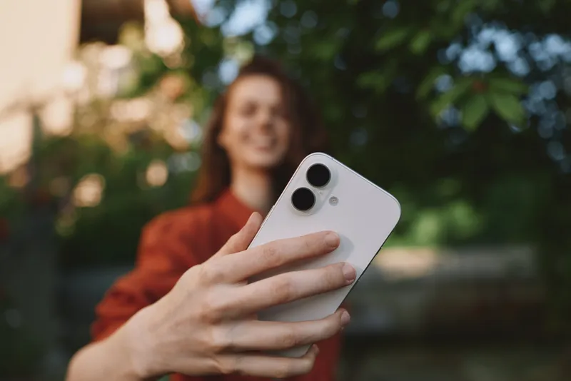 Young woman in casual sweater takes a sunny selfie outdoors, smiling softly as lush greenery surrounds the candid moment