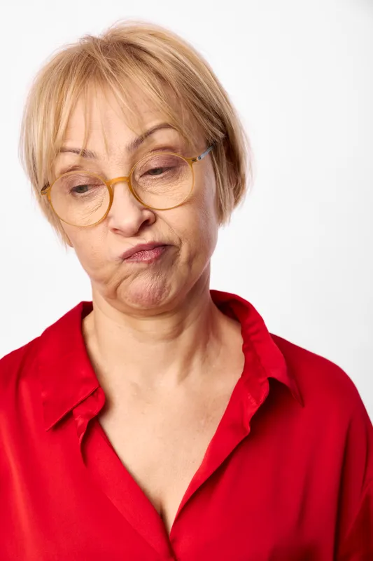 Woman elderly blonde glasses portrait red shirt indoor closeup on white background, she looks down with thoughtful expression, soft light, quiet calm posture