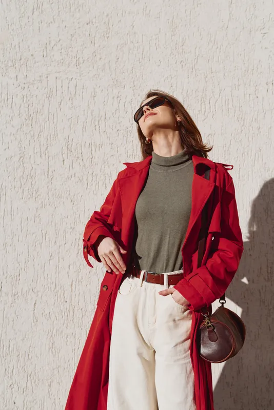 Elegant woman wearing a red trench coat over a gray top and white jeans stands against a pale textured wall, sunglasses on, looking upward with confidence and a stylish urban mood.