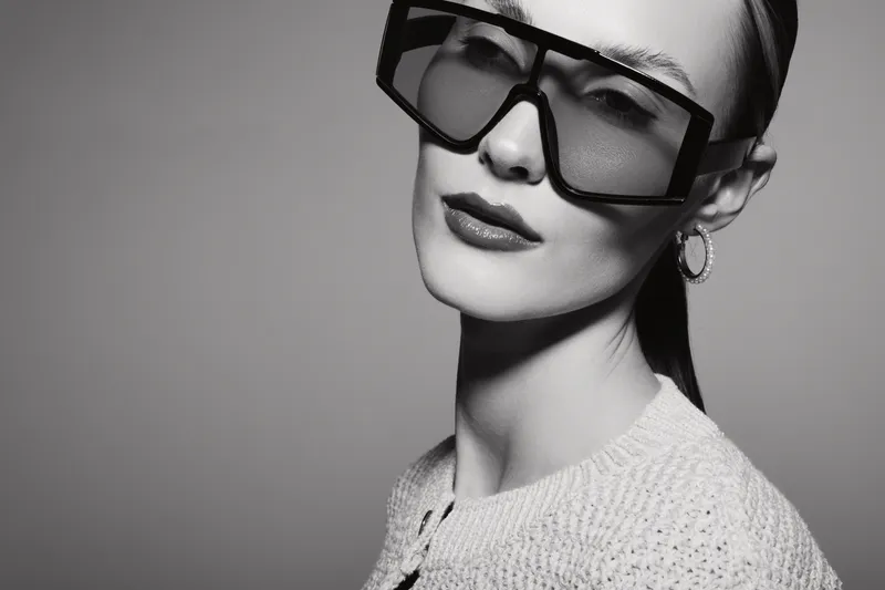 Stylish white woman with pale skin wears large square sunglasses and textured sweater. Studio portrait in black and white with high contrast lighting and minimalistic background.