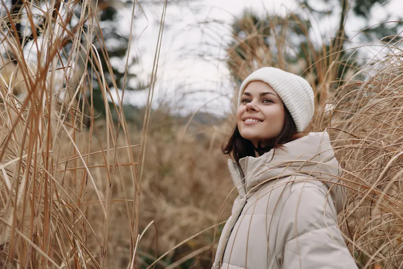 Woman wearing a beige coat and white beanie stands in tall grasses, smiling in a serene outdoor winter portrait with soft natural light