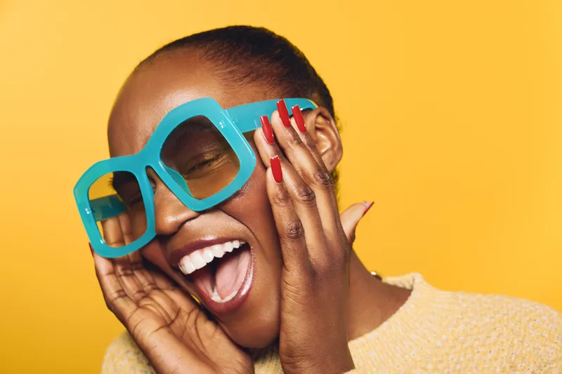 Happy African American woman with dark skin wearing large blue sunglasses and red nail polish smiles with eyes closed on yellow background. Concept of joy, fashion, and summer style.