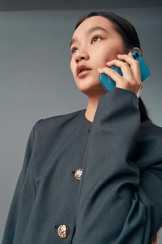 Young woman with long hair wearing dark blazer talking on smartphone looking sideways. Communication, technology, casual portrait with modern device and thoughtful expression on face.