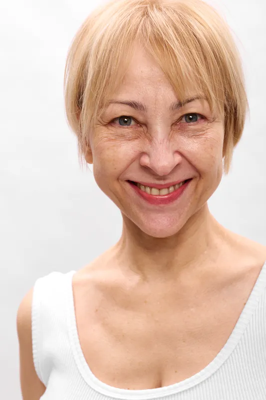 Woman portrait smiling elderly blonde closeup studio white top calm friendly expression, soft studio light, visible wrinkles, relaxed shoulders, neutral background, natural beauty and wellness