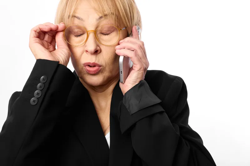 Businesswoman in black suit, wearing glasses, holding a phone to her ear, eyes closed with headache expression, stress at work, communication call, isolated on white background, soft studio light,