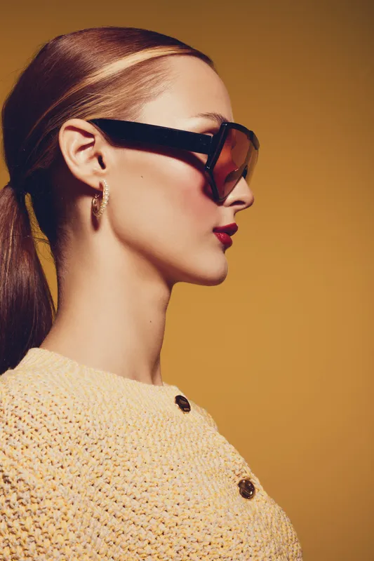 Profile of a stylish woman with sunglasses and red lipstick wearing a knitted sweater against a yellow background. Concept of fashion, beauty, and modern style.