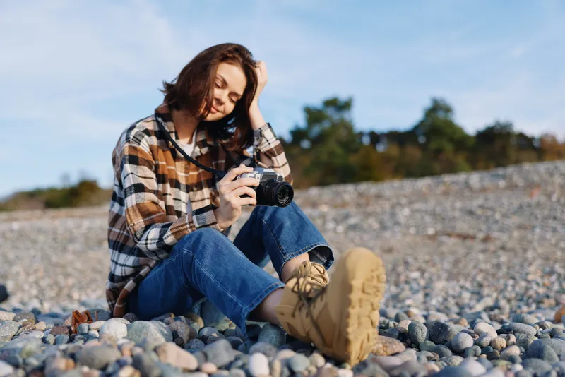 Woman photographer on a pebbled beach, wearing a plaid shirt and boots, concentrates on her camera as waves retreat. Natural light highlights textures, a calm outdoor portrait with serene coastal