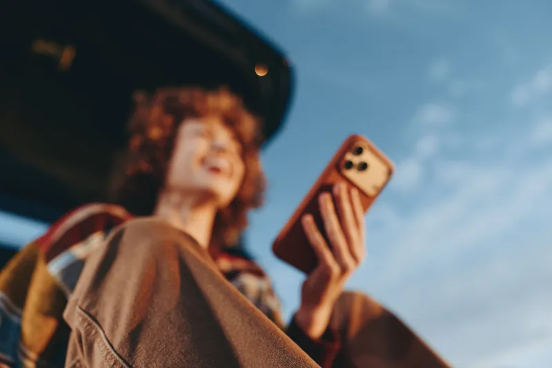 Woman with curly hair takes a selfie outdoors, holding a smartphone up against a bright sky, casual lifestyle moment, soft sunlight, candid expression for daily communication