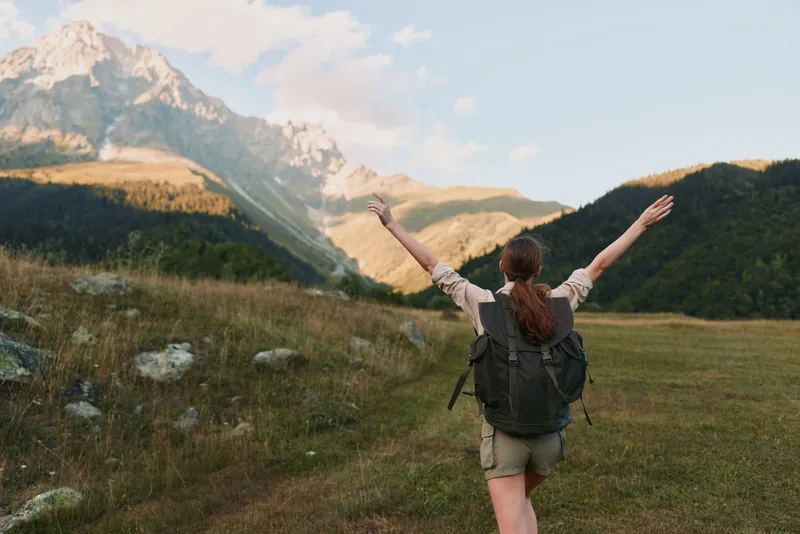 Cheerful woman outdoors, arms raised in triumph, backpacked hiker enjoying alpine meadow with mountains in soft light, vibrant greens, and a sense of freedom, breeze.