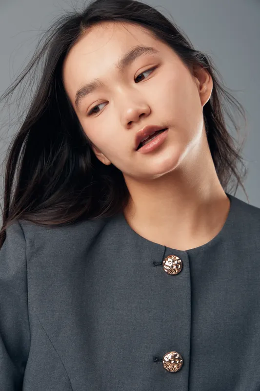 Portrait of young woman with long black hair wearing gray buttoned coat looking down with neutral expression on gray background for fashion and beauty concept.