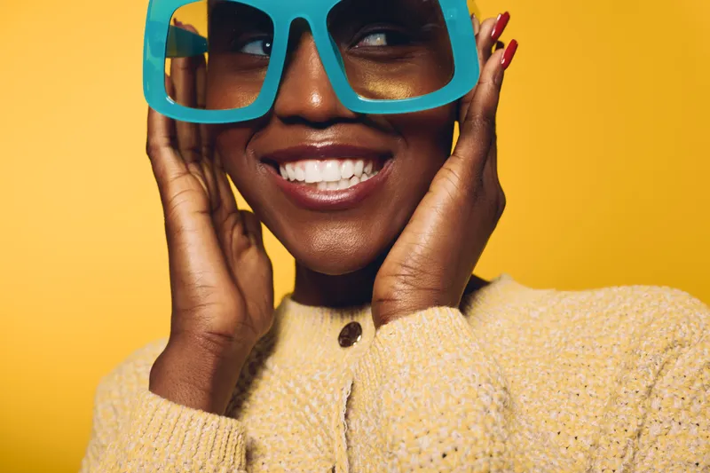 Smiling dark skinned African woman wearing large blue sunglasses and a beige sweater poses against yellow background. Concept of fashion, joy, and vibrant summer style.
