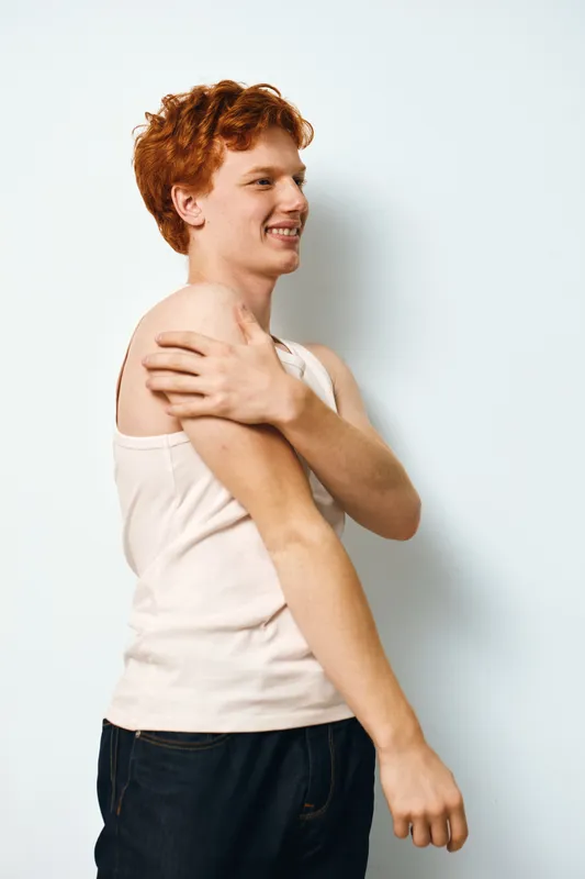 Young man with red hair wearing sleeveless white shirt standing against white background smiling and touching shoulder with hand in casual pose and relaxed mood.