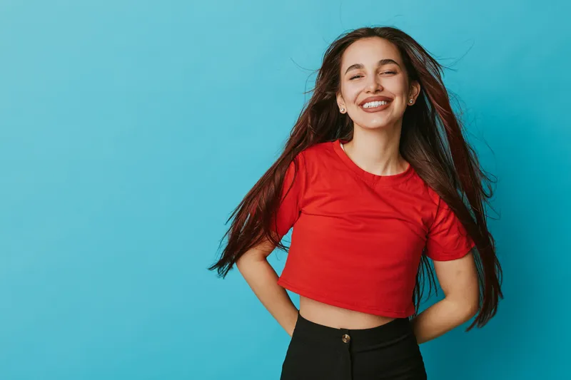Young woman smiling with long hair wearing red crop top and black pants standing against blue background with hands behind back and eyes closed in joyful expression.