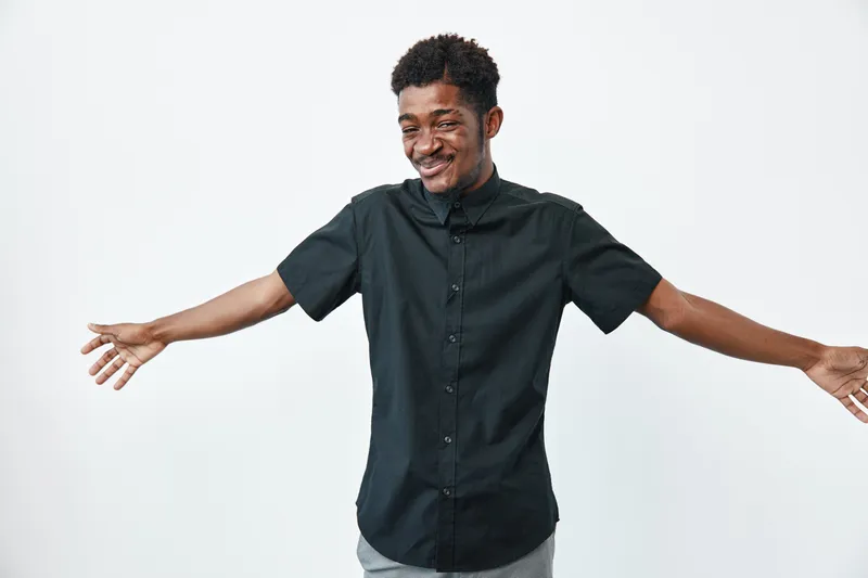 Young black man with dark skin wearing black shirt standing with arms wide open smiling on white background. Male adult, casual style and happy expression for positive emotion.