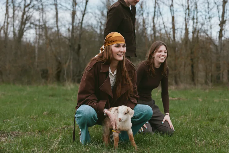 Young woman in a brown jacket and yellow headscarf crouches outdoors holding a baby goat while two friends smile nearby in a grassy field during a casual lifestyle moment.
