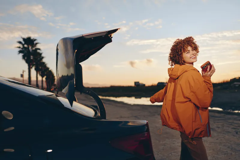 Woman lifestyle outdoors at sunset, standing near open car trunk by ocean, wearing orange jacket, holding drink cup, soft wind, seaside travel break, warm golden light, relaxed smile