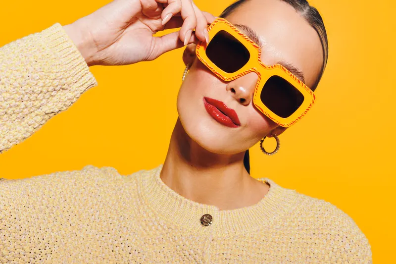 Stylish woman in yellow sunglasses and beige sweater poses confidently against bright yellow background. Fashion concept with bold colors and modern accessories for trendy look.