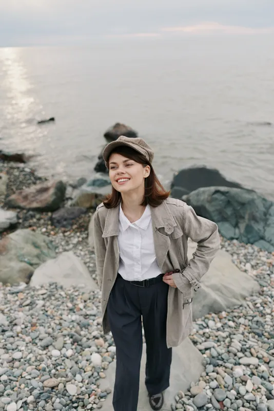 Young woman in casual clothes stands on rocky beach near calm sea, smiling and looking away. Concept of leisure, nature, casual fashion, and outdoor lifestyle by the water.