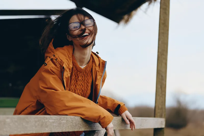 Woman lifestyle outdoors, smiling portrait in orange jacket and glasses, leaning on wooden railing, warm daylight, relaxed pose, soft umbrella shade, calm mood, natural background