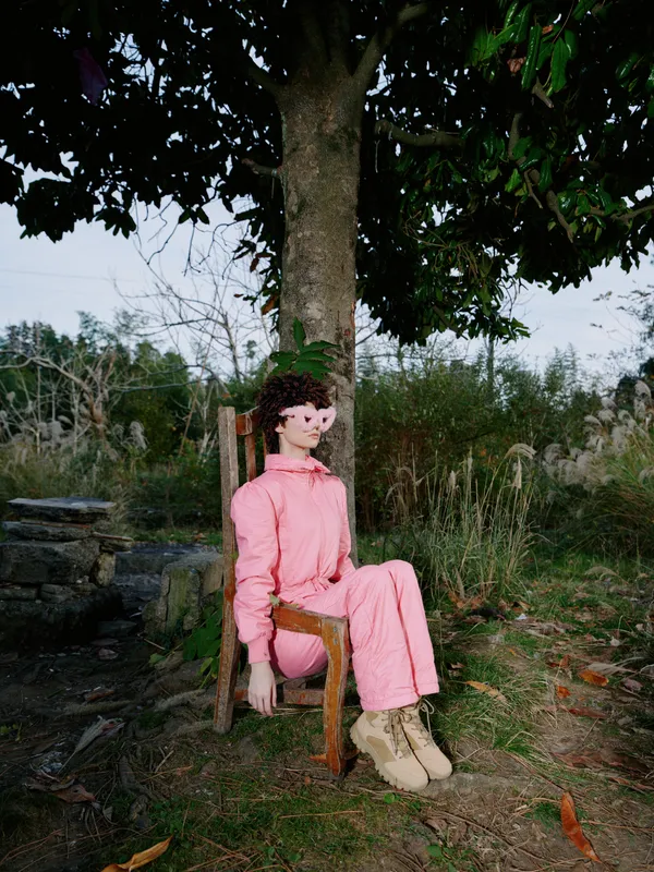 Woman in pink outfit sits on a wooden chair under a tree in a rustic outdoor setting, calm portrait with natural background and soft light for garden or rural lifestyle
