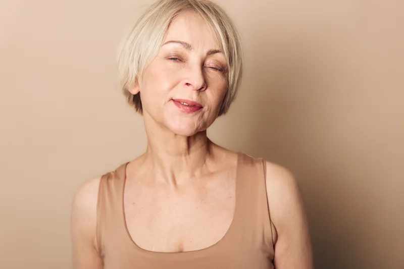 Blonde middle aged woman with short hair and closed eyes smiles gently wearing beige tank top. Neutral beige background creates calm and natural mood for skincare or wellness concept.