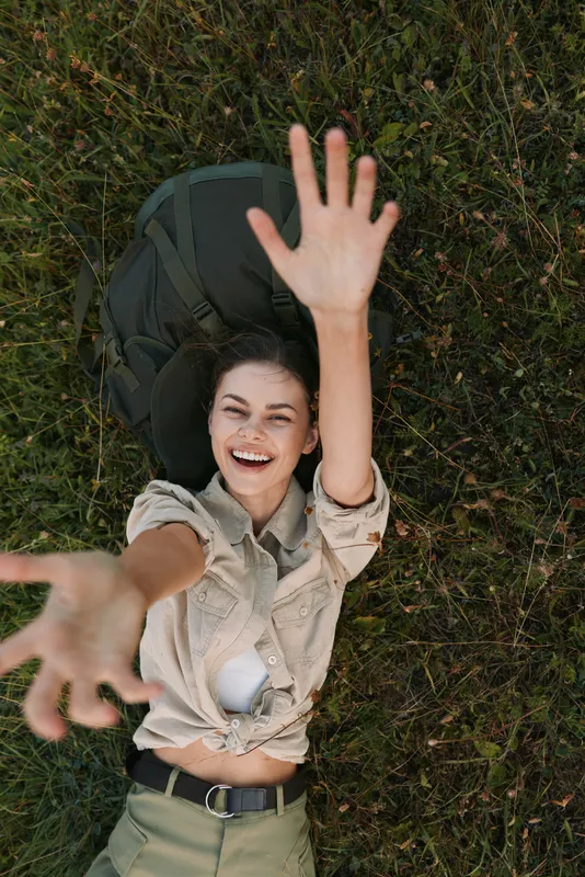 A happy young woman lies on grass reaching her hands towards the camera smiling. Outdoor lifestyle concept with natural light and casual clothing in a relaxed pose.