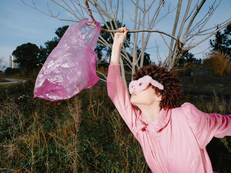 Woman in pink sweater outdoors, reaching toward a pink plastic bag hung on a tree branch, playful pose against a natural landscape of grass and bare trees at dusk.