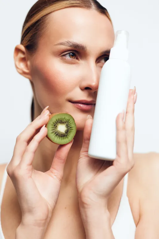 Woman beauty skincare routine, holding kiwi beside her cheek while showing a cosmetic bottle, concept of fresh fruit ingredient for hydration, gentle facial care, studio purity and selfcare today