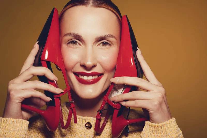 Smiling woman holds red high heel shoes near face, wearing yellow sweater. Studio portrait with warm lighting on brown background. Concept of fashion, style, and happiness.