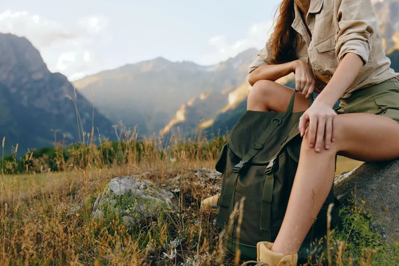 Cheerful woman outdoors, seated on a rock with a backpack, enjoying sunlit mountain landscape, relaxed lifestyle moment, natural colors, fresh air and horizon