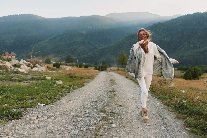 Cheerful woman outdoors on a gravel road, white outfit, flowing cardigan, sunlit countryside, mountains in background, relaxed lifestyle moment and breeze soft color tone