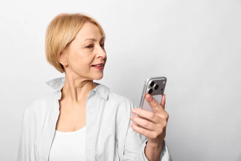 Senior woman holding smartphone, smile while viewing screen, relaxed portrait in casual white blouse, communication and technology theme, isolated on white background