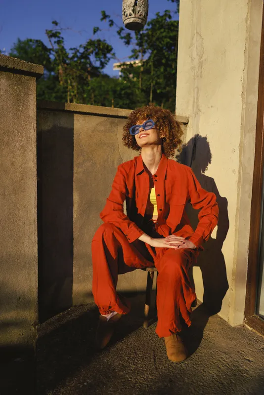 Cheerful woman outdoors in a red suit, relaxed pose, sunlit rooftop setting, curly hair, sunglasses, fashion lifestyle portrait with warm natural tones and greenery in the background