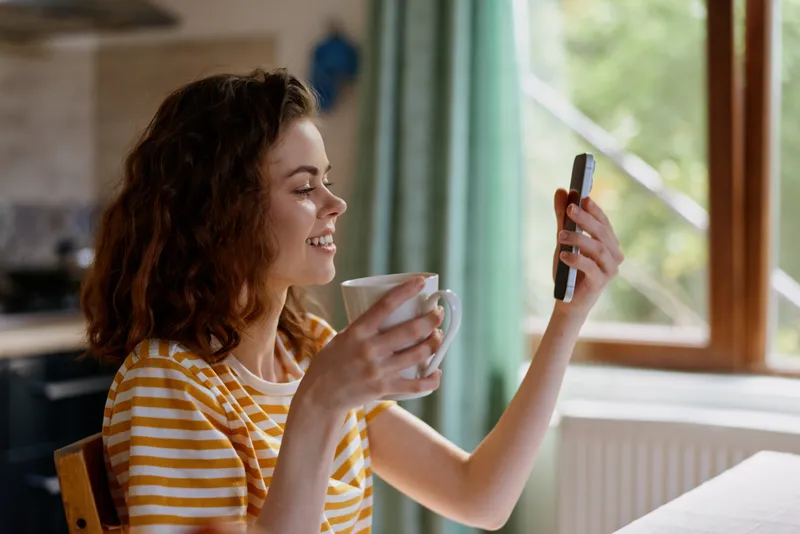Woman in striped shirt enjoys a warm morning at home, sipping coffee while taking a selfie, smiling at her phone by a sunlit window, creating a cozy lifestyle moment